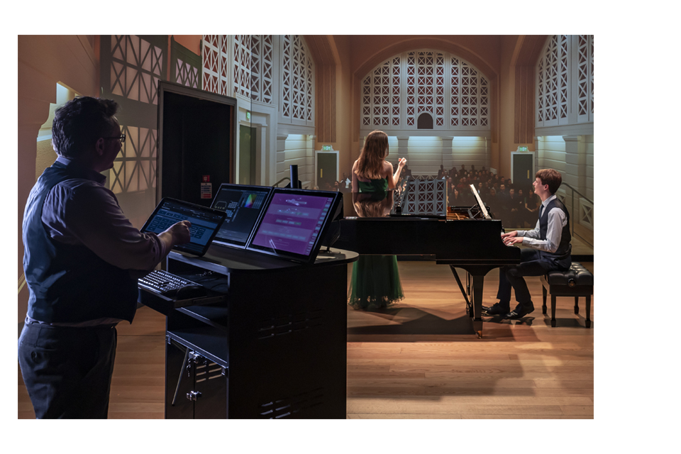 A man wearing a light blue shirt, using digital tablet to control a visual simulator, with two students, a female soprano and a male pianist, performing in front of a simulator.